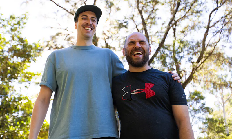 Tony and his Hireup support worker David are standing side by side in a park, with an arm wrapped tightly across each other's shoulders. Sunlight seeps through the leafy trees. Tony is bald, has a beard and wears a navy top. David has a moustache, wears a cap and light blue top.