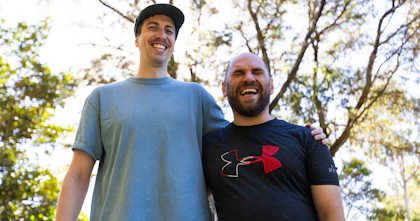 Tony and his Hireup support worker David are standing side by side in a park, with an arm wrapped tightly across each other's shoulders. Sunlight seeps through the leafy trees. Tony is bald, has a beard and wears a navy top. David has a moustache, wears a cap and light blue top.