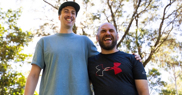 Tony and his Hireup support worker David are standing side by side in a park, with an arm wrapped tightly across each other's shoulders. Sunlight seeps through the leafy trees. Tony is bald, has a beard and wears a navy top. David has a moustache, wears a cap and light blue top.
