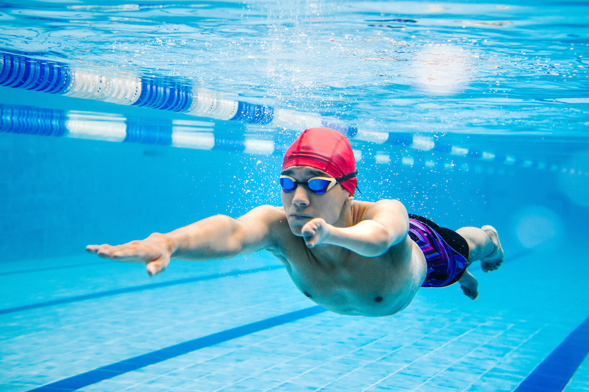 A young man is swimming in a lap pool. He wears a red cap and goggles. 