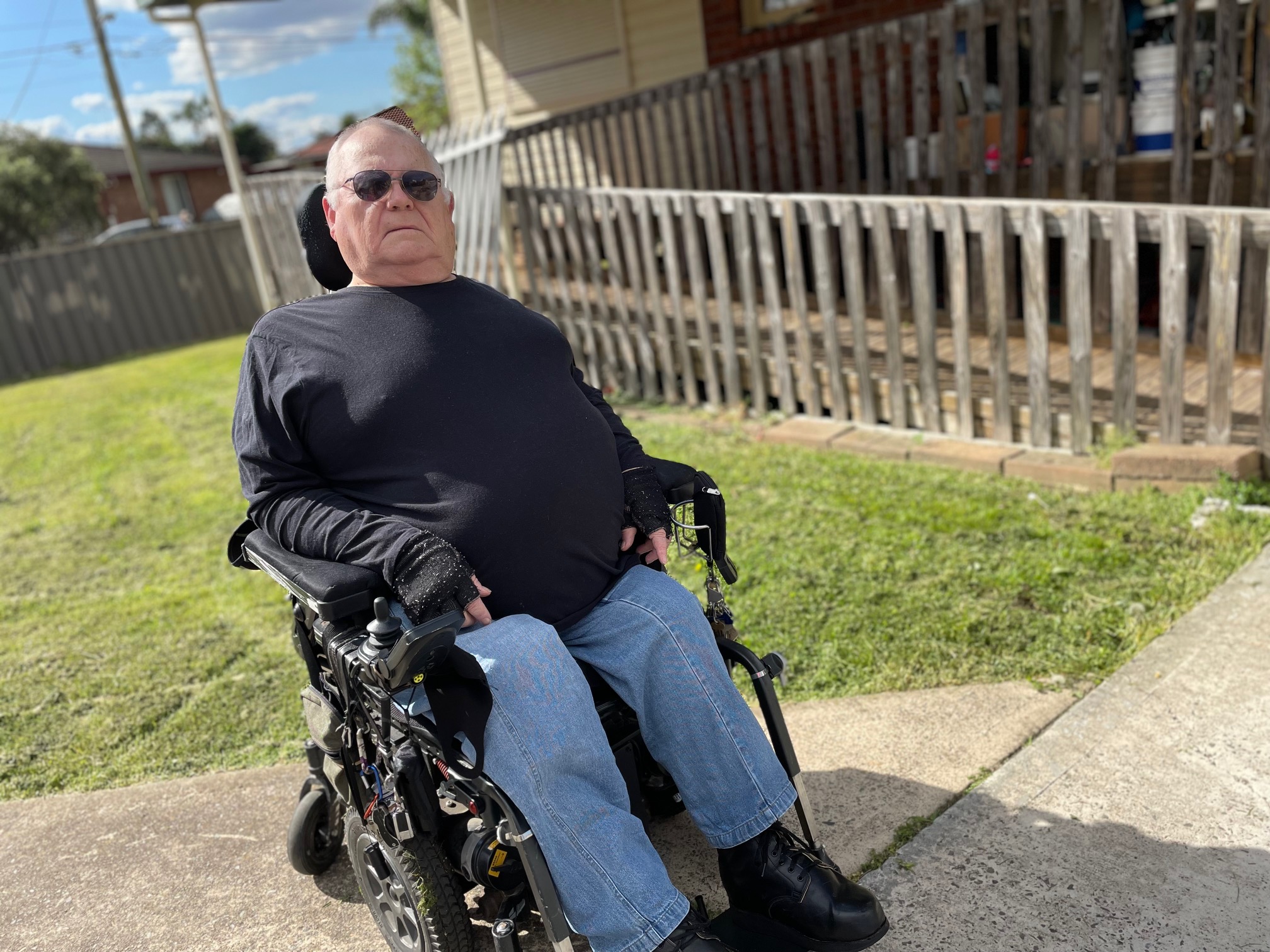 George Laszuk uses a wheelchair. He is on a footpath wearing a black long sleeved shirt and blue jeans. There is grass and wooden fence in the background.