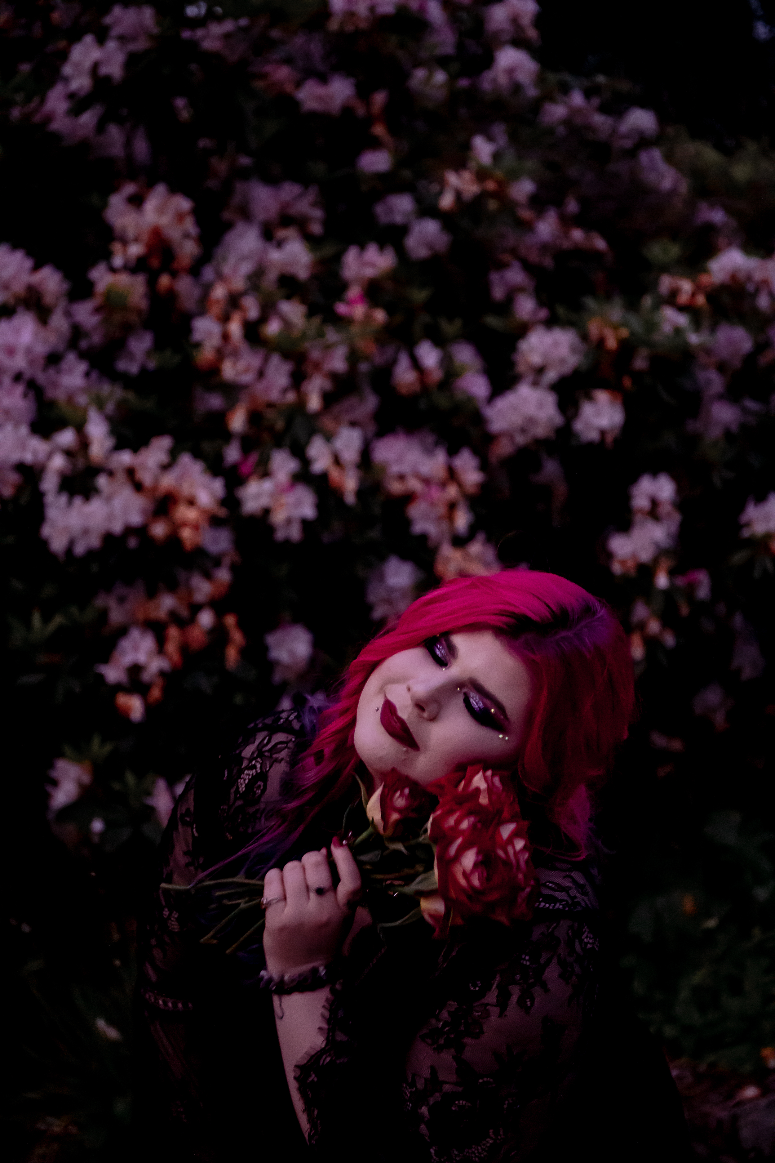 Disability advocate Zoe Simmons has long pink hair and wears a black lace dress. She stands against a floral backdrop while holding a bunch of roses. Photo by Emma Veness