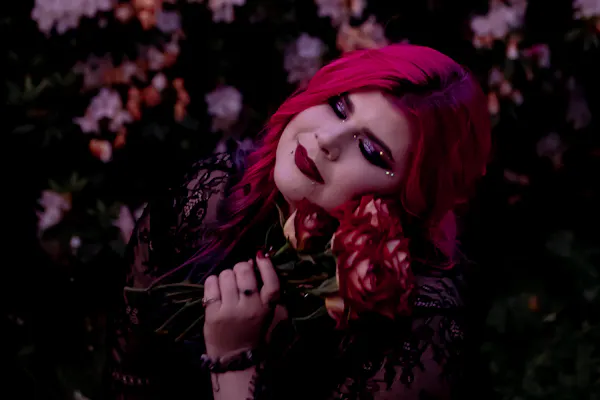 Disability advocate Zoe Simmons has long pink hair and wears a black lace dress. She stands against a floral backdrop while holding a bunch of roses. Photo by Emma Veness