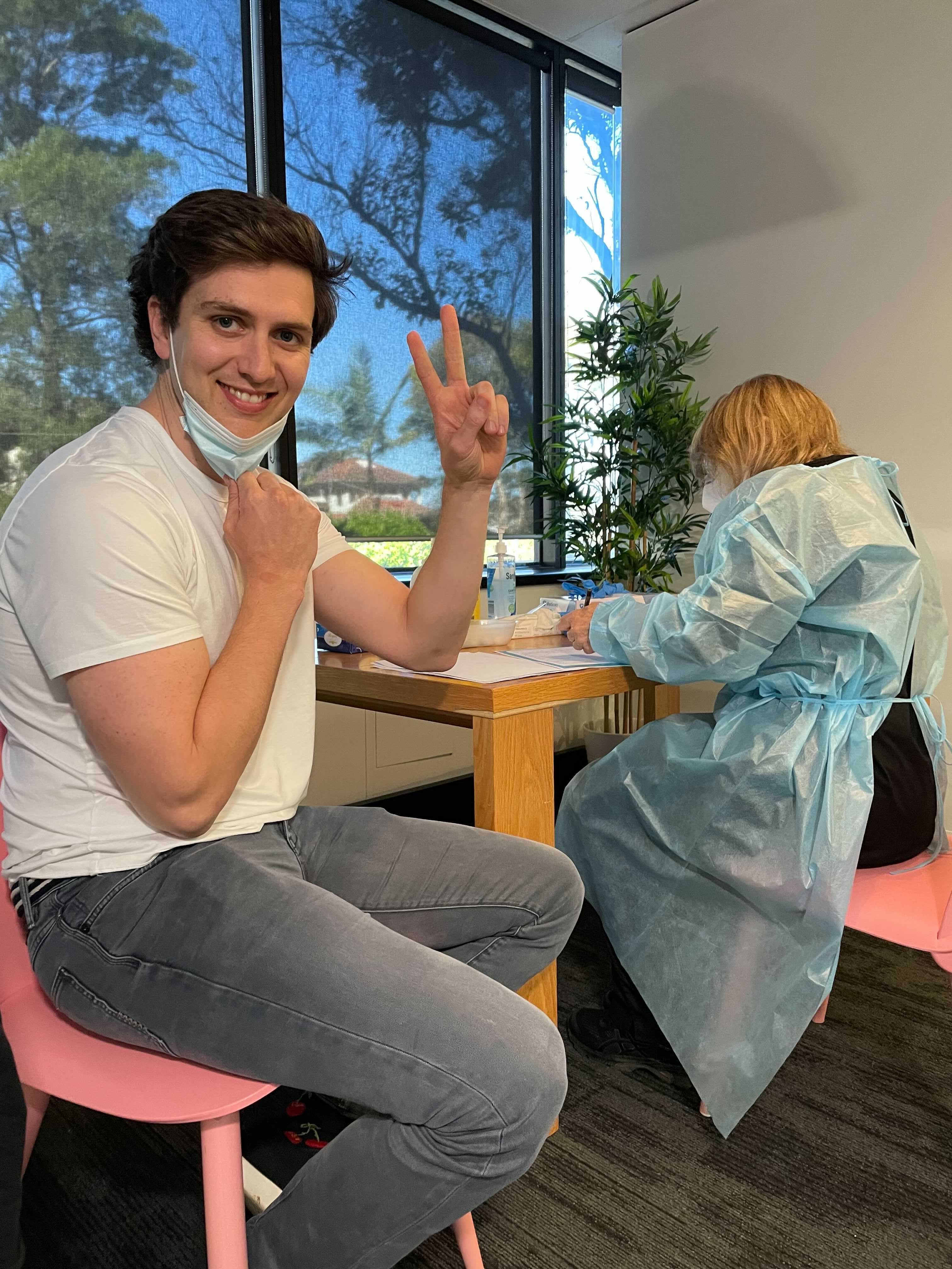 Hireup Co-Founder and CEO Jordan O'Reilly preparing to be vaccinated. He sits on a pink chair and is doing the peace sign with his fingers while smiling at the camera. A nurse sits at a desk in the background. 
