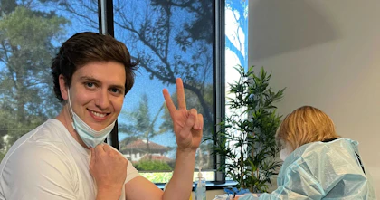 Hireup Co-Founder and CEO Jordan O'Reilly preparing to be vaccinated. He sits on a pink chair and is doing the peace sign with his fingers while smiling at the camera. A nurse sits at a desk in the background.