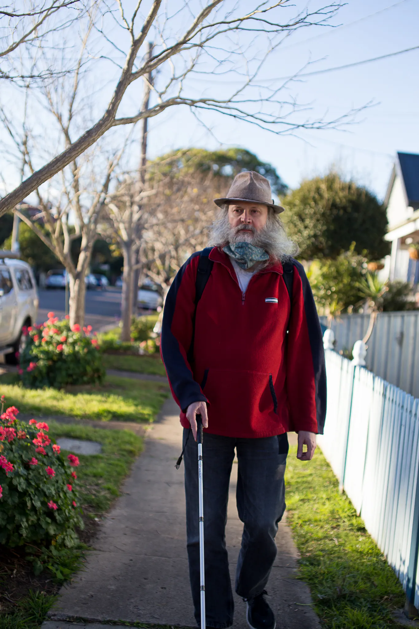 Andrew, a man with a large white beard is wearing a hat. He is walking down a street with houses on one side and trees on the other. He is using a cane.
