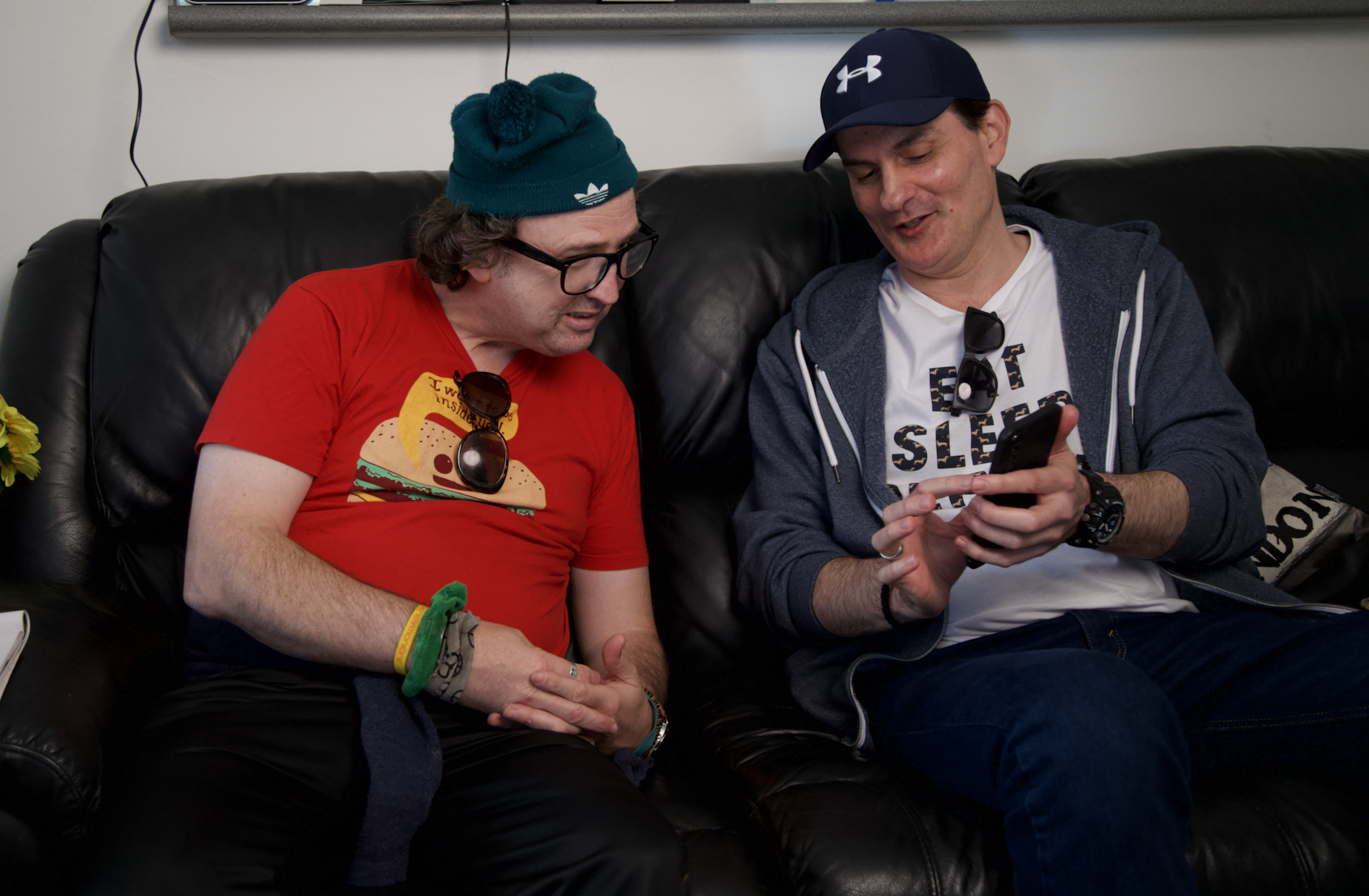 David and Goran are sitting together on a leather couch and Goran is showing David something on his phone. David is wearing a green beaning and a red t-shirt. Goran is wearing a white t-shirt, blue hoodie and blue cap.