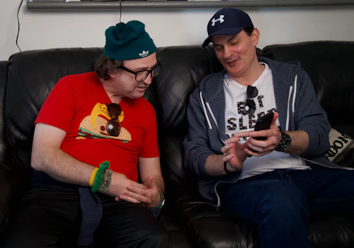 David and Goran are sitting together on a leather couch and Goran is showing David something on his phone. David is wearing a green beaning and a red t-shirt. Goran is wearing a white t-shirt, blue hoodie and blue cap.