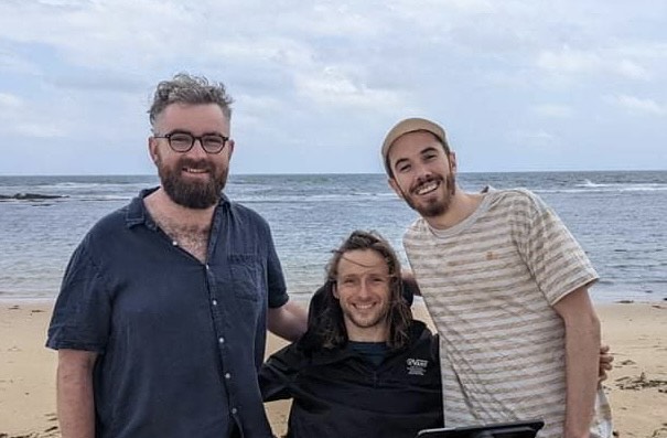 Steve is with two friends on the beach. They have their arms around each other and are smiling at the camera. 
