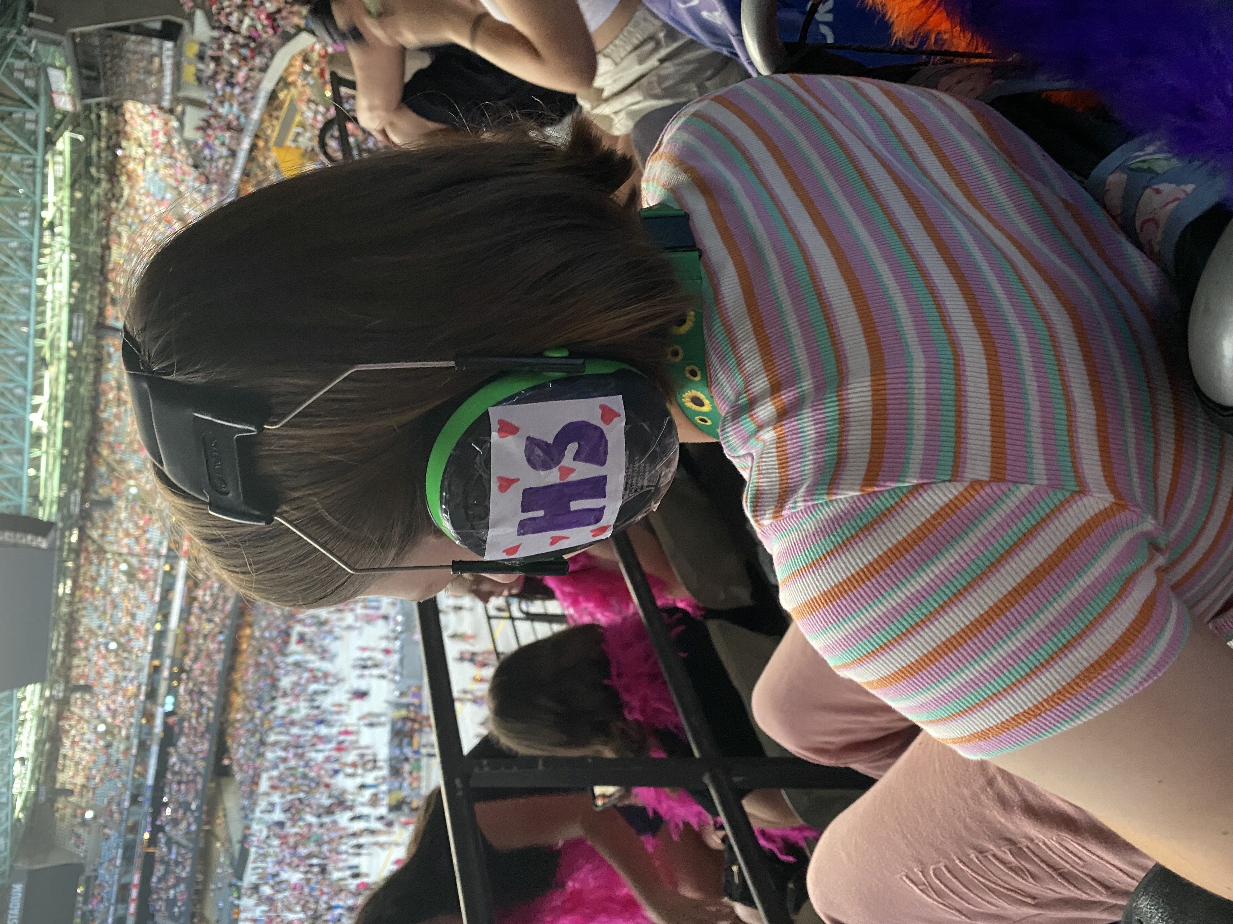 A teenager wearing noise-cancelling headphones and a colourful striped t-shirt sits in the crowd at a music concert.