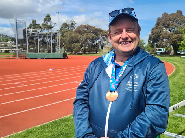 Hireup client Caleb is wearing a Special Olympics jacket and gold medal. He's standing beside a running track.