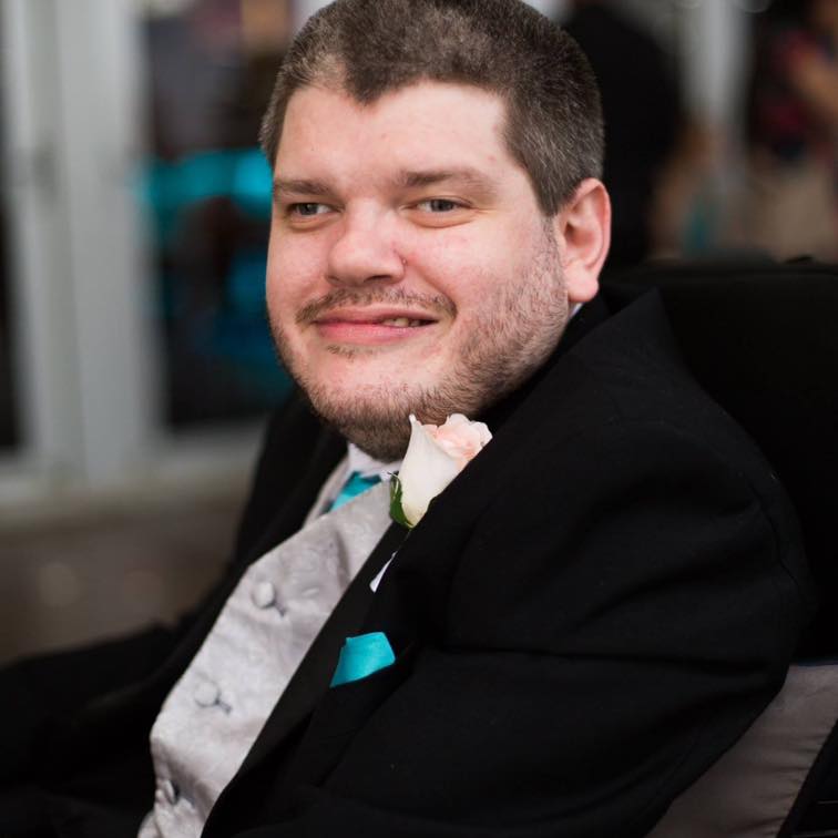 Disability activist Todd Winther smiles while attending a formal event. He wears a black suit jacket, silver suit vest, white shirt, aqua blue tie and pocket square, and a light pink rose boutonniere. 