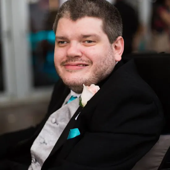 Disability activist Todd Winther smiles while attending a formal event. He wears a black suit jacket, silver suit vest, white shirt, aqua blue tie and pocket square, and a light pink rose boutonniere.
