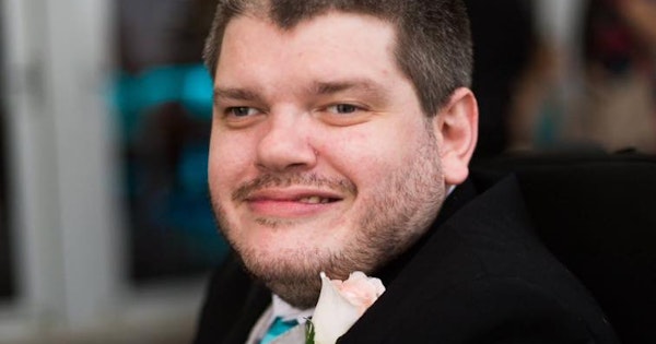 Disability activist Todd Winther smiles while attending a formal event. He wears a black suit jacket, silver suit vest, white shirt, aqua blue tie and pocket square, and a light pink rose boutonniere.