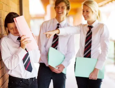 A school boy and girl are pointing and laughing at another school girl who is holding up her book to protect herself.