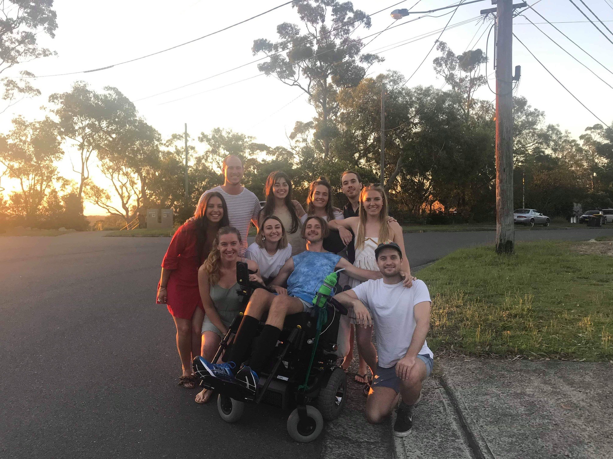 Steven is pictured with a group of friends who are standing around him. They're on a street corner and the sun is shining through eucalyptus trees.
