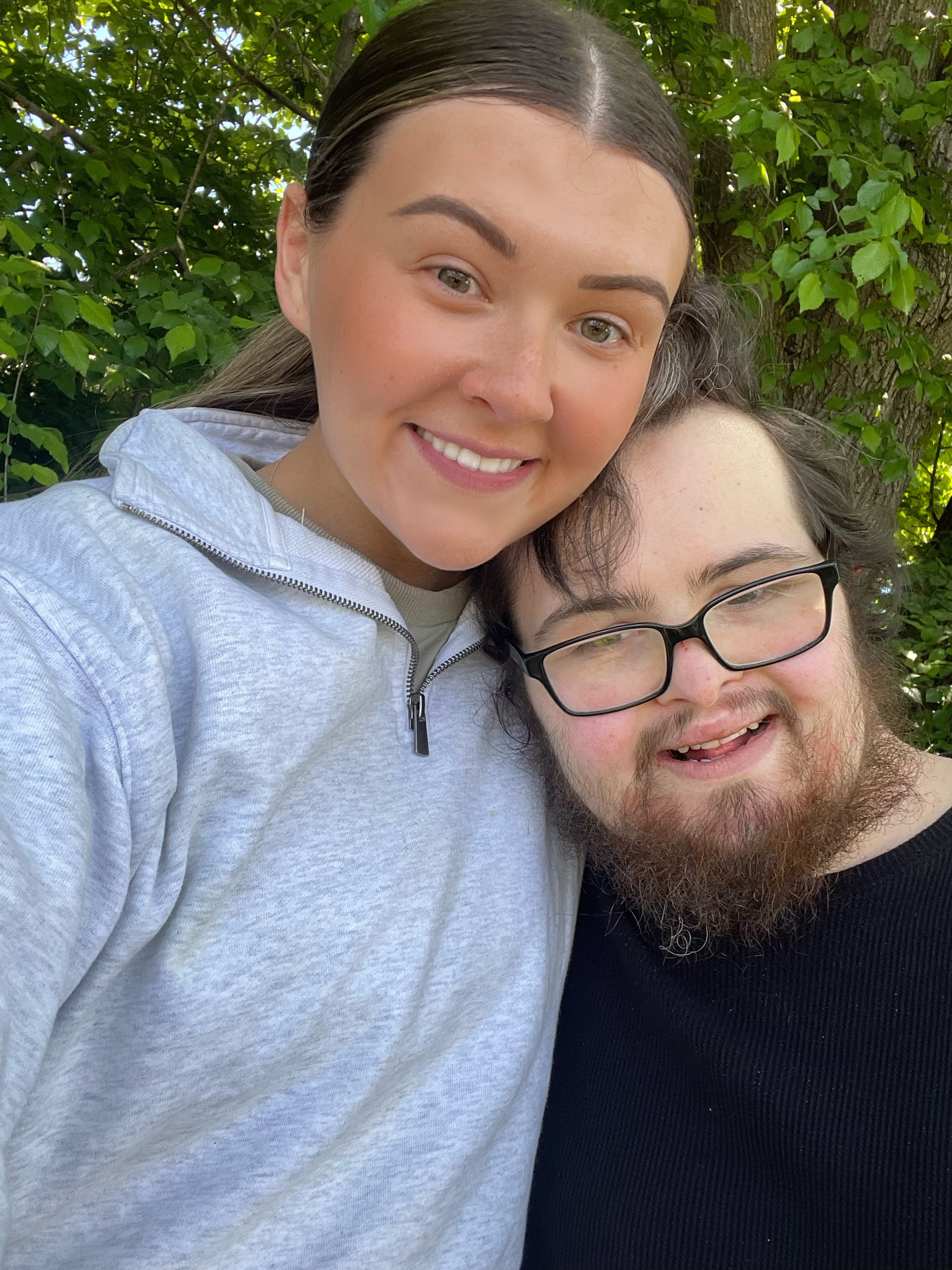 Ebony and Riley are smiling together. Ebony has long brown hair and is wearing a light grey sports hoodie. Riley has brown hair, a beard and is wearing reading glasses and a black singlet.