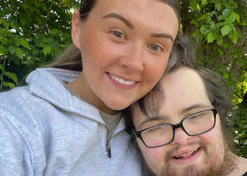 Ebony and Riley are smiling together. Ebony has long brown hair and is wearing a light grey sports hoodie. Riley has brown hair, a beard and is wearing reading glasses and a black singlet.