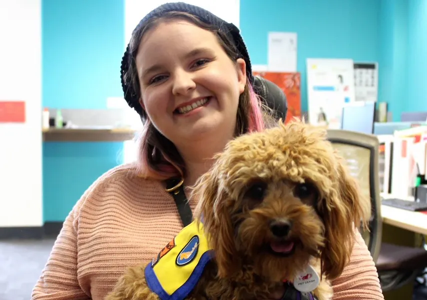 Jess has a light brown dog on her lap. The dog is wearing a yellow and blue harness. Jess has pink hair and is wearing a salmon coloured jumper. She is smiling and holding the dog. Jess uses a wheelchair.