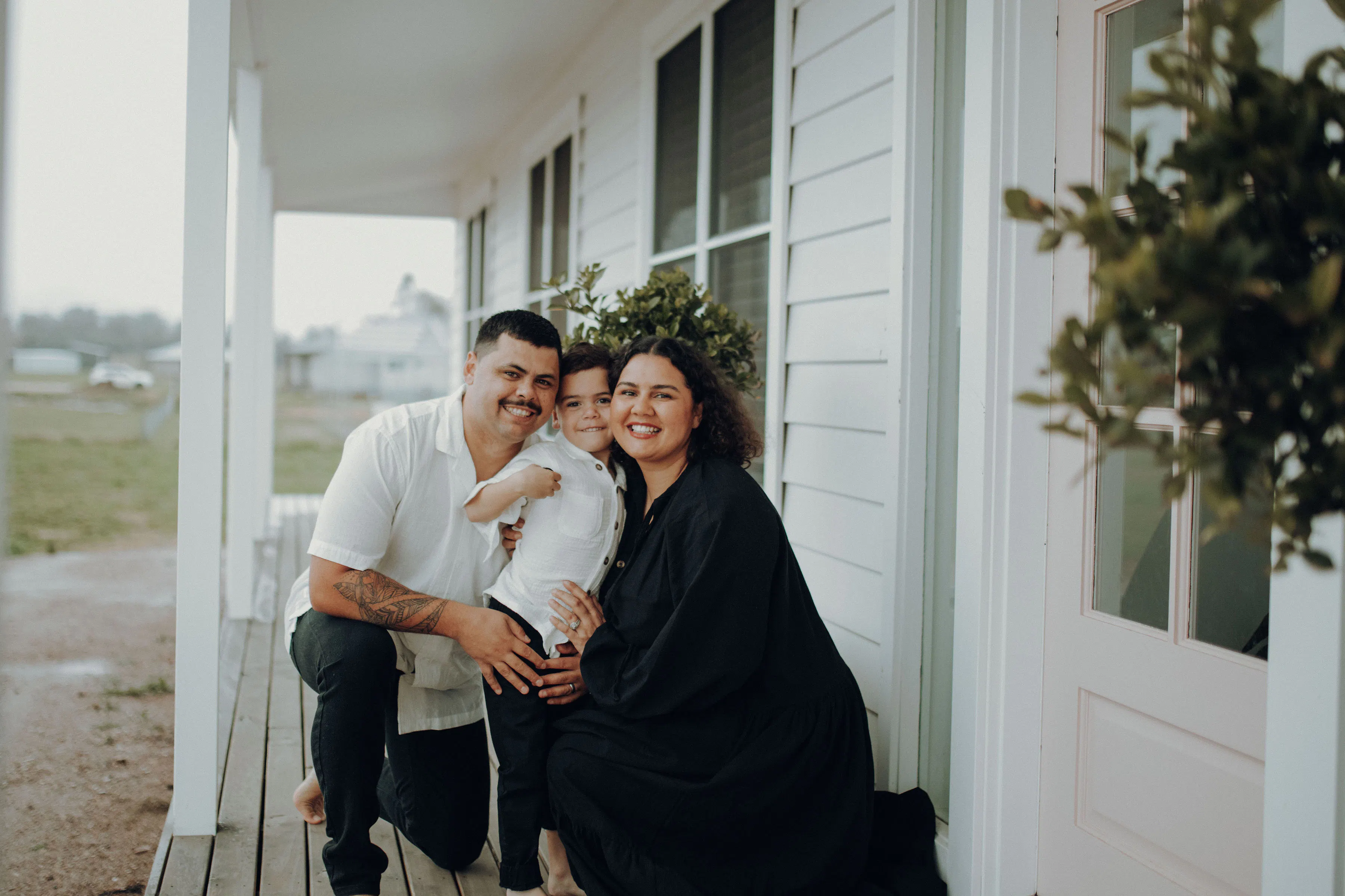 A smiling father, young son and mother huddle together on the porch of a house.