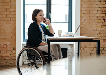 A woman with short brown hair is sitting at a desk using a mobile phone. A laptop, paper cup, pens and some white headphones are on the desk in front of her. She is wearing a black blazer, white top and cream long pants. The woman is a wheelchair user.