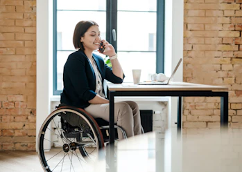 A woman with short brown hair is sitting at a desk using a mobile phone. A laptop, paper cup, pens and some white headphones are on the desk in front of her. She is wearing a black blazer, white top and cream long pants. The woman is a wheelchair user.