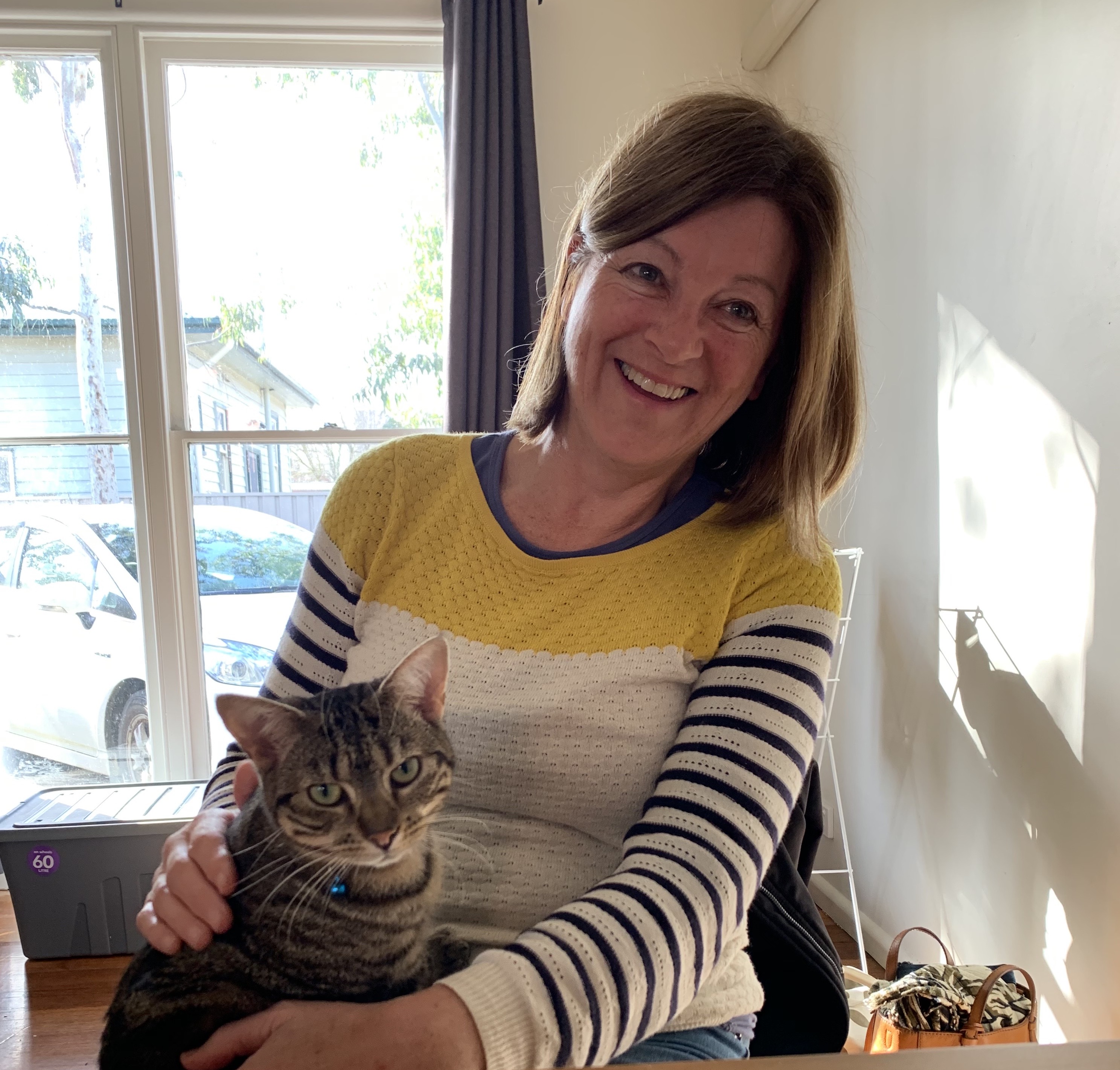 Annette is wearing a striped jumper and is patting a cat that is sitting peacefully on Annette's lap. 