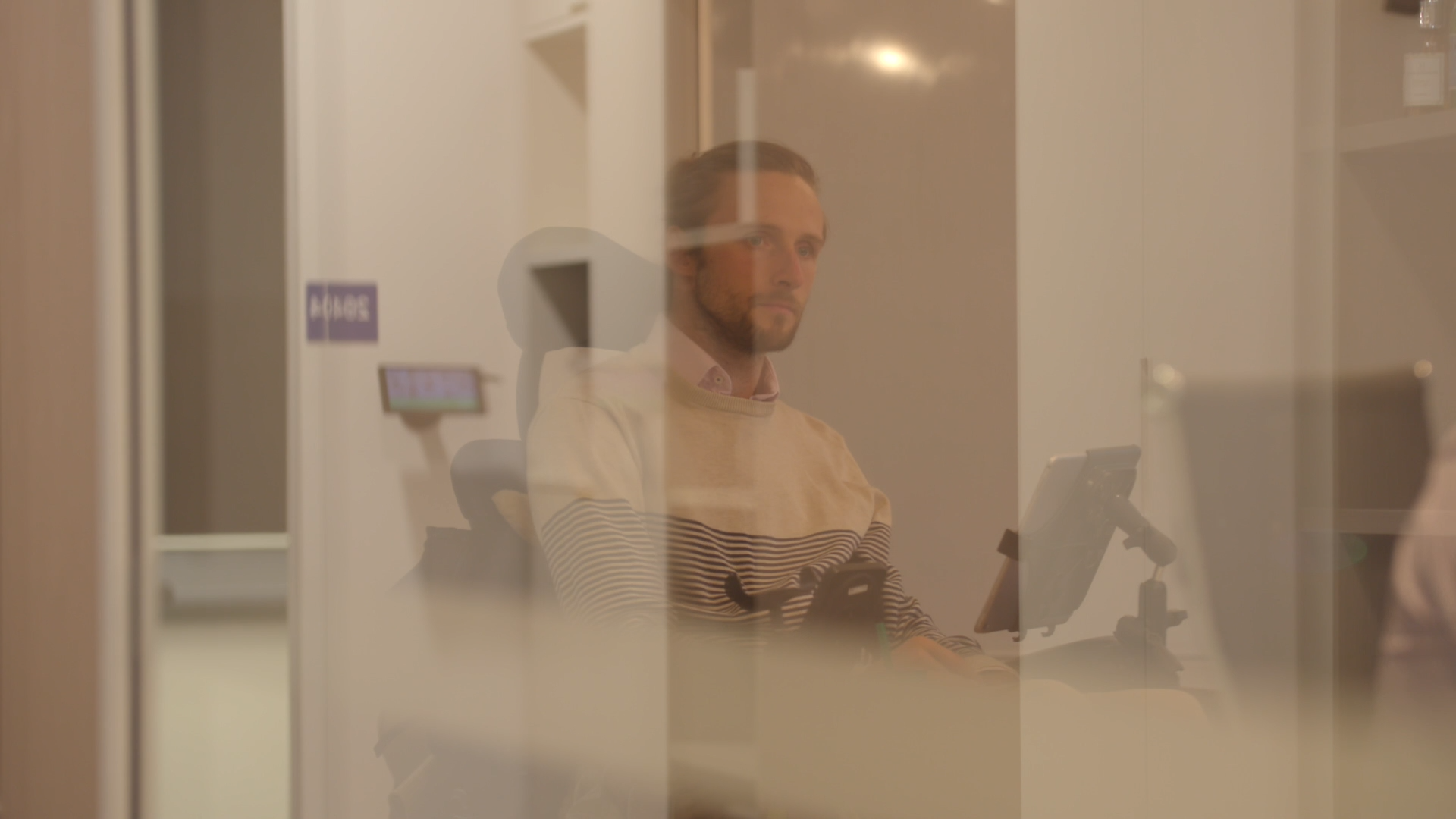 Steve Ralph is sitting on the other side of a window. Steve uses a wheelchair and is in a meeting room. He has long hair that is tied back.