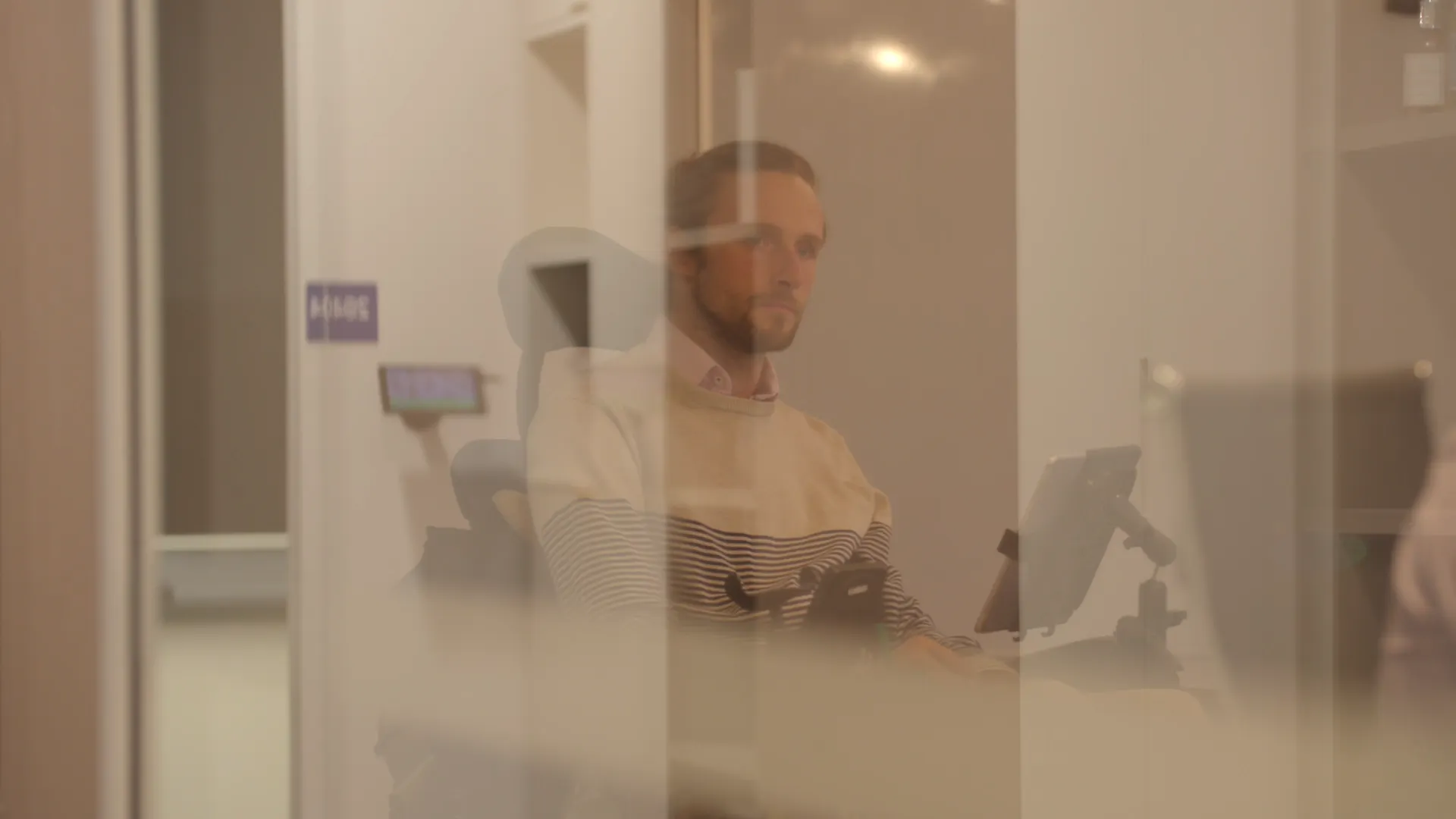Steve Ralph is sitting on the other side of a window. Steve uses a wheelchair and is in a meeting room. He has long hair that is tied back.