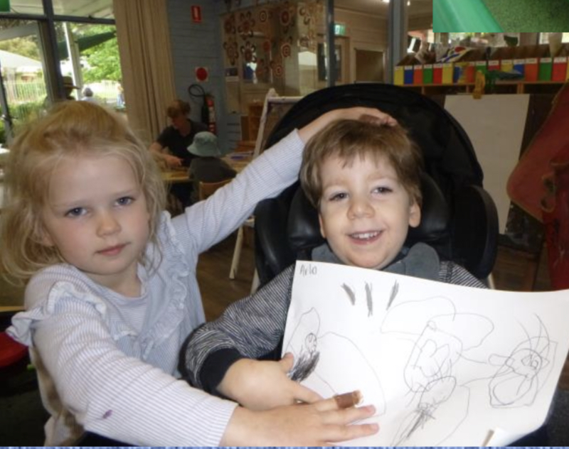 A boy and girl sit together smiling at their preschool, holding up a drawing.