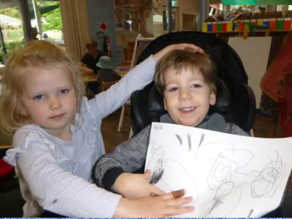 A boy and girl sit together smiling at their preschool, holding up a drawing.