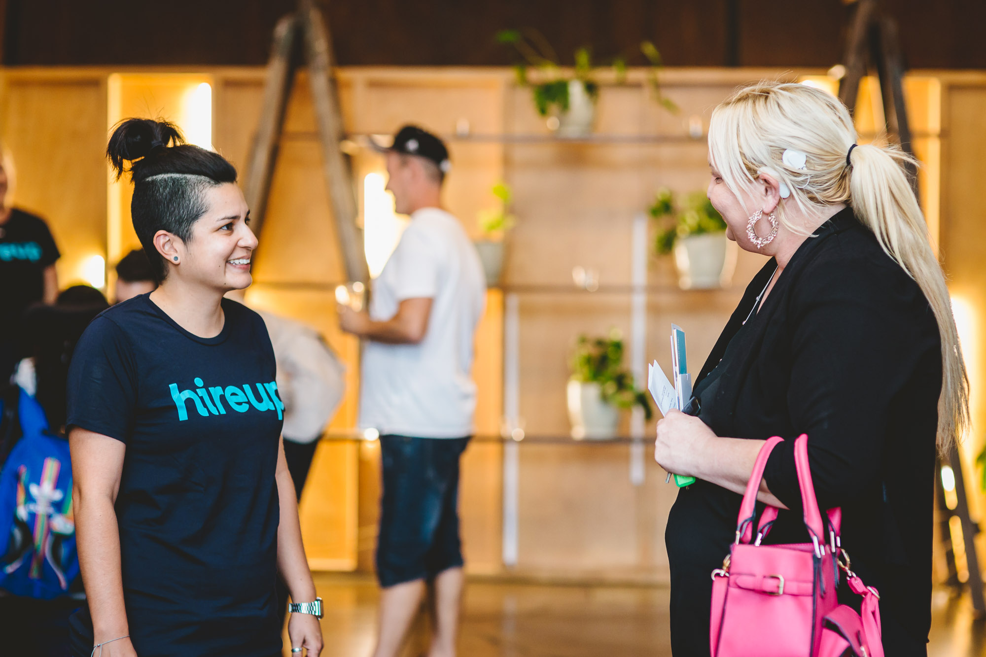 A woman with black hair tied up wears a shirt that says 'Hireup' on it. Standing next to her is a woman with blonde hair who is holding pamphlets and a pink handbag. She has a Cochlear implant. 