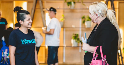 A woman with black hair tied up wears a shirt that says 'Hireup' on it. Standing next to her is a woman with blonde hair who is holding pamphlets and a pink handbag. She has a Cochlear implant.