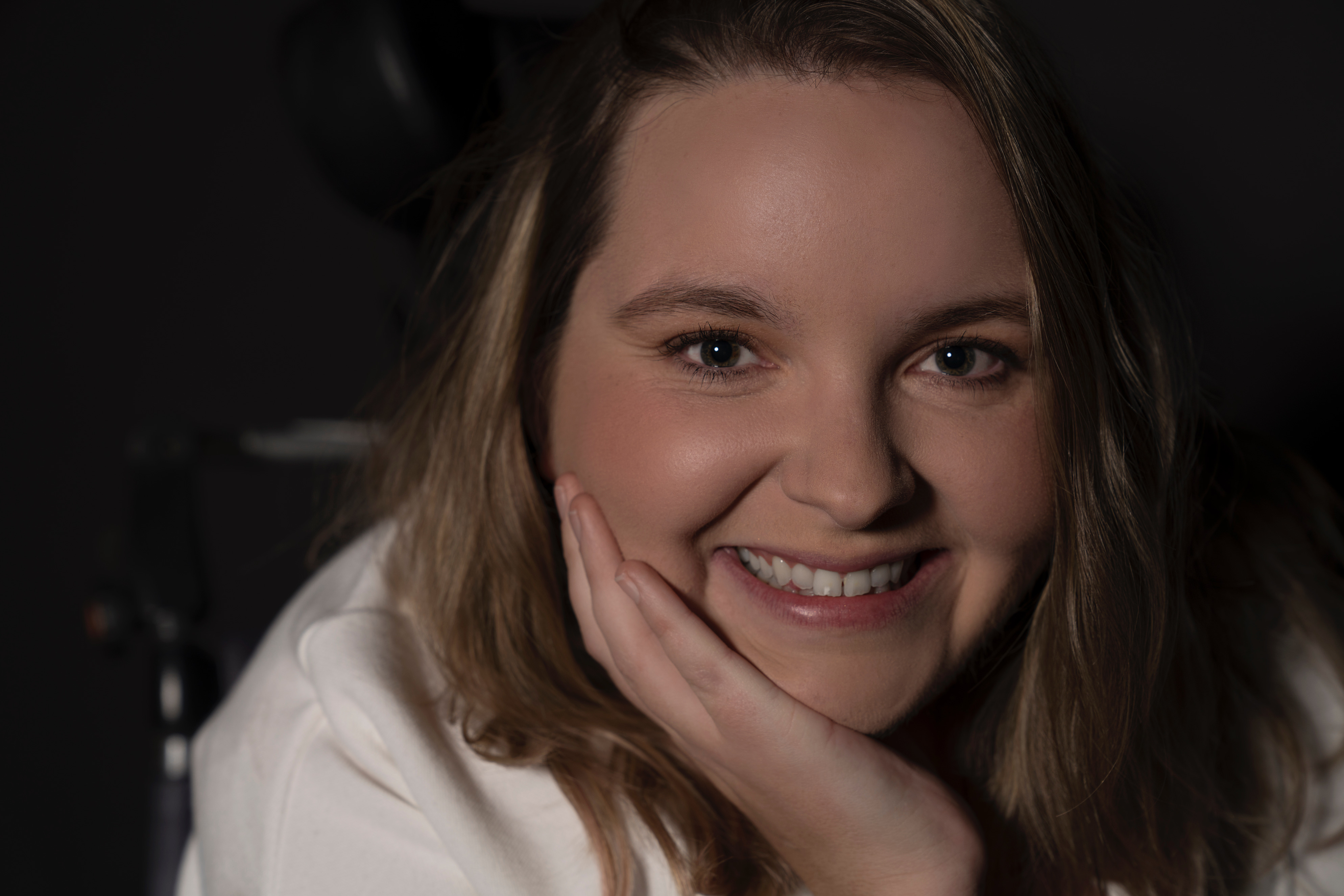Jess is smiling with her hand on her cheek. She has light brown hair. Jess is a wheelchair user.