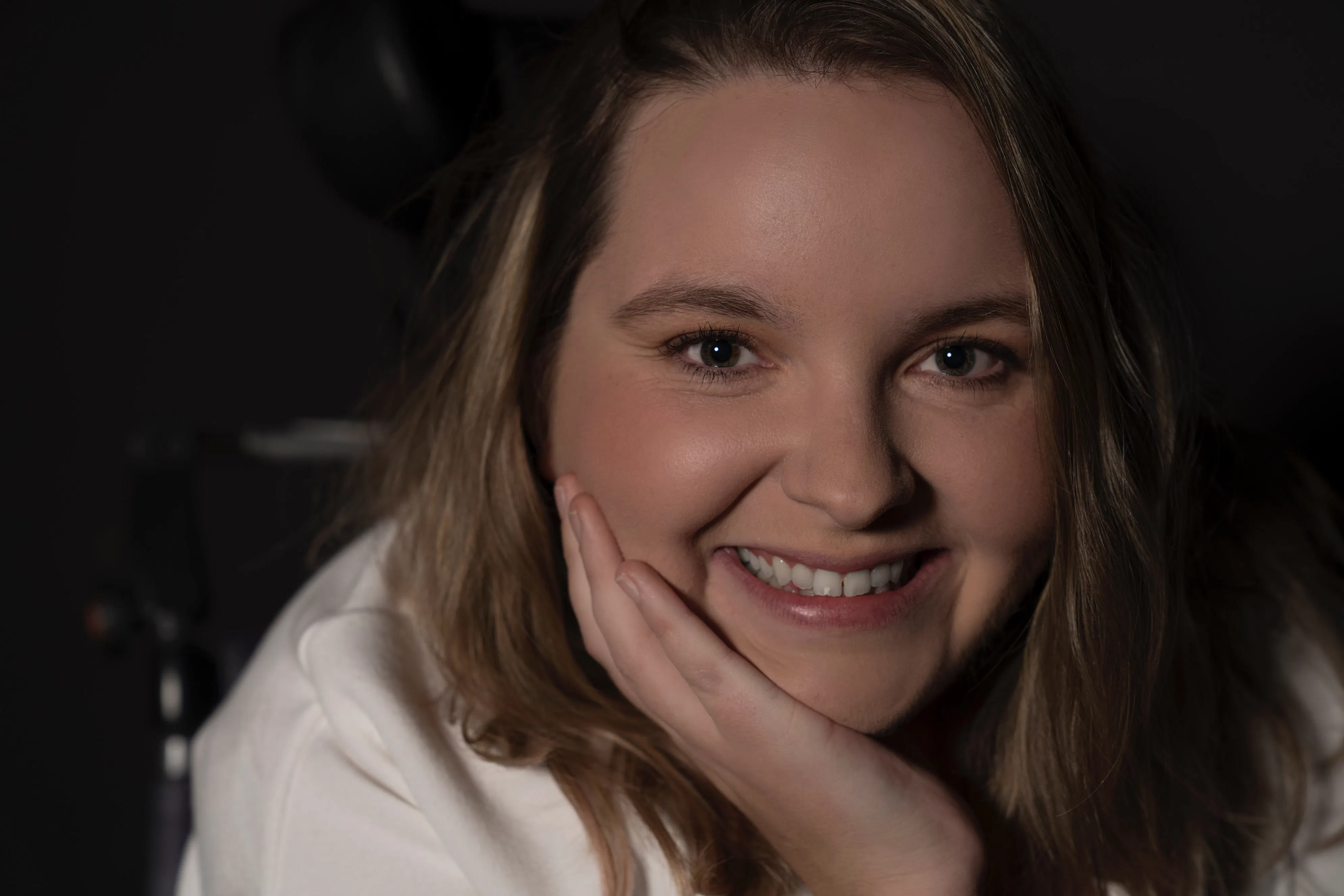 Jess is smiling with her hand on her cheek. She has light brown hair. Jess is a wheelchair user.