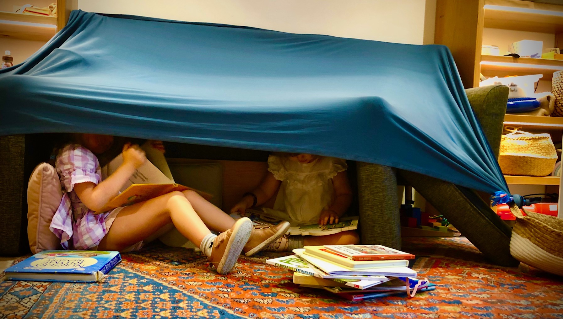 Two children read books under a fort made from a sheet and couch cushions. They are surrounded by piles of books and baskets filled with toys. 