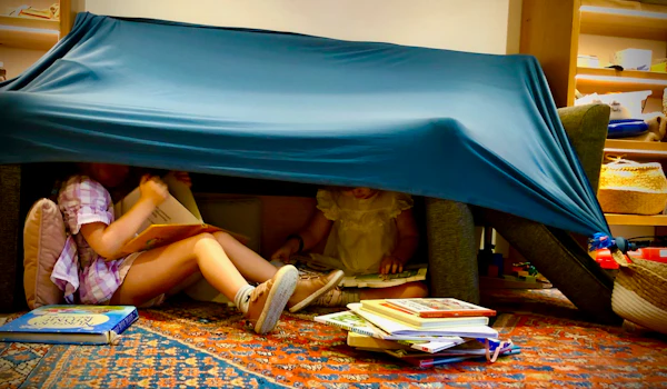 Two children read books under a fort made from a sheet and couch cushions. They are surrounded by piles of books and baskets filled with toys.