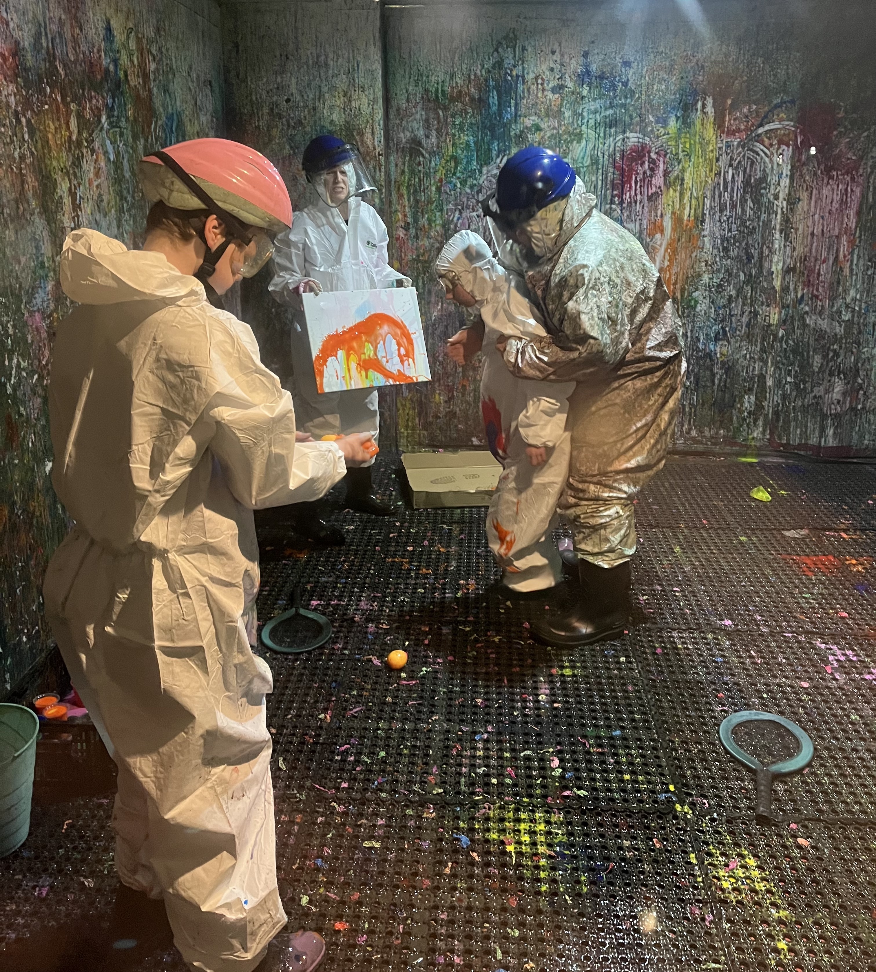 A group people wearing protective helmets and white jumpsuits are in a 'splash room' covered in paint. One holds a canvas that the other throw paint at. 