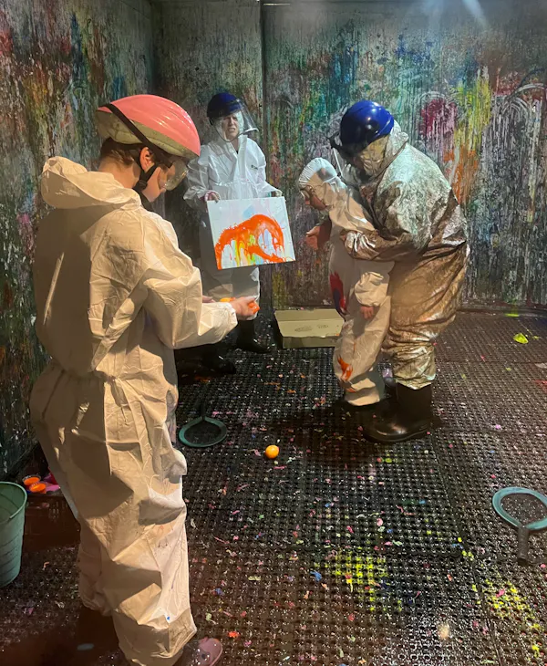 A group people wearing protective helmets and white jumpsuits are in a 'splash room' covered in paint. One holds a canvas that the other throw paint at.