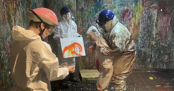 A group people wearing protective helmets and white jumpsuits are in a 'splash room' covered in paint. One holds a canvas that the other throw paint at.