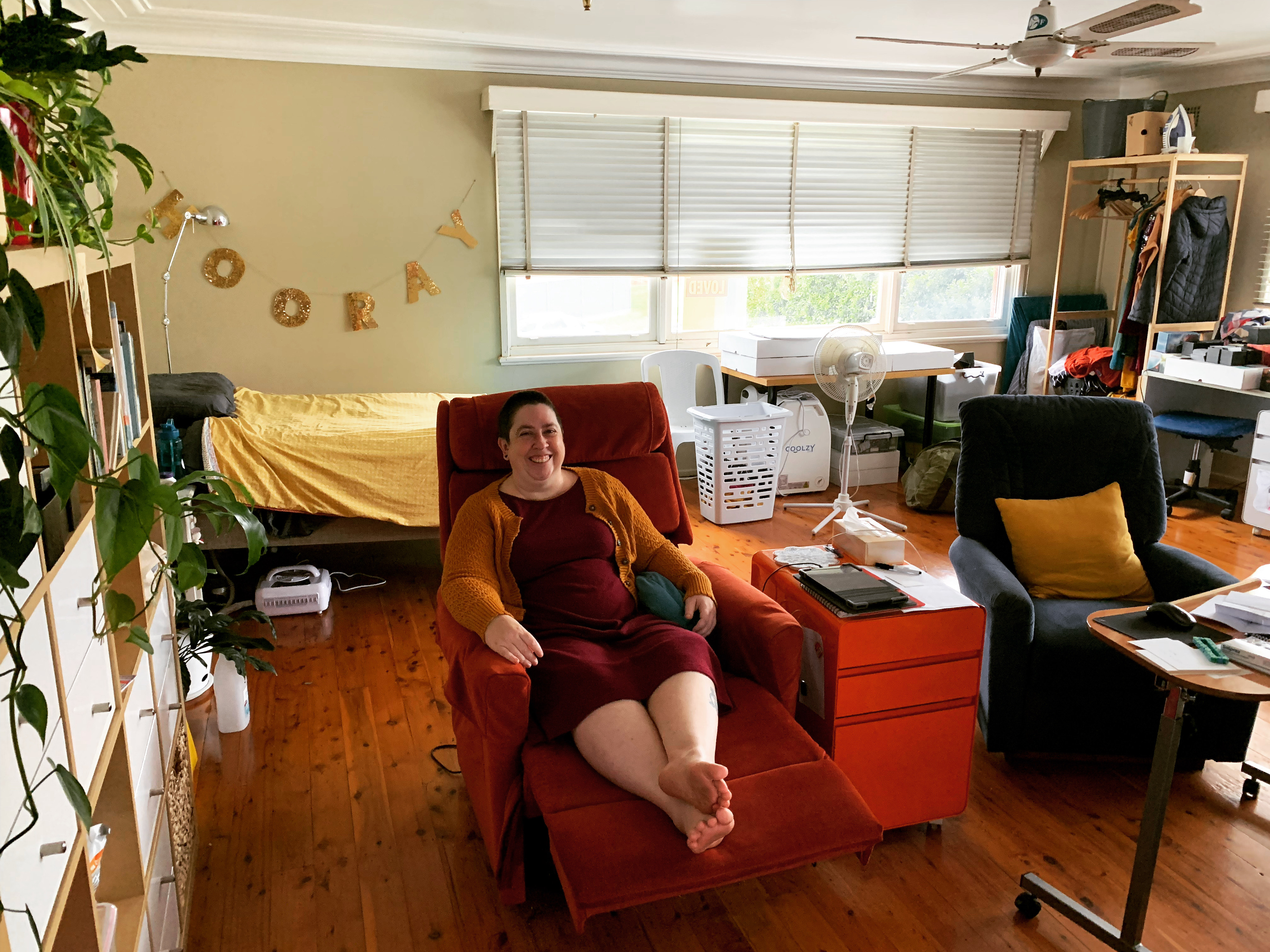 Catherine is sitting in a laid back sofa chair and smiling at the camera. There is a plant in the foreground, a sign that says 'Hooray' and a window in the background, and a couch and table on the right hand side. 