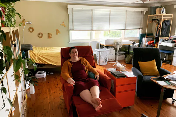 Catherine is sitting in a laid back sofa chair and smiling at the camera. There is a plant in the foreground, a sign that says 'Hooray' and a window in the background, and a couch and table on the right hand side.