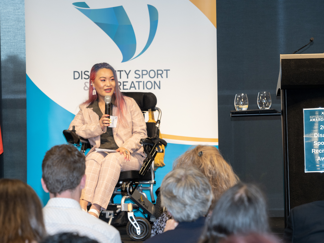 Akii presents to an audience. They're holding a microphone and a banner behind them reads: Disability Sport and Recreation. Akii is a wheelchair user.
