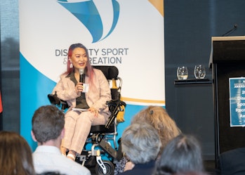 Akii presents to an audience. They're holding a microphone and a banner behind them reads: Disability Sport and Recreation. Akii is a wheelchair user.