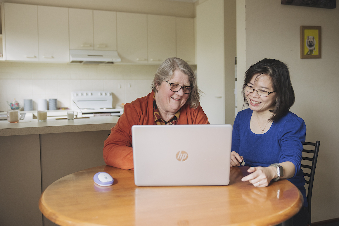 Hireup client Leanne and her support worker Dieu are seated in front of a laptop screen. They're both looking at the laptop and are smiling. 