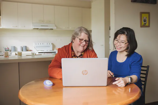 Hireup client Leanne and her support worker Dieu are seated in front of a laptop screen. They're both looking at the laptop and are smiling. 