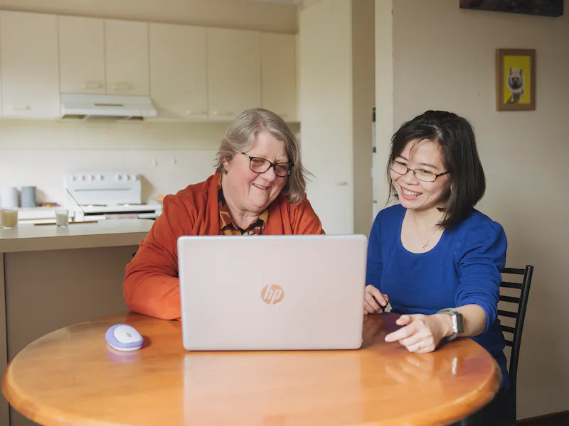 Hireup client Leanne and her support worker Dieu are seated in front of a laptop screen. They're both looking at the laptop and are smiling.