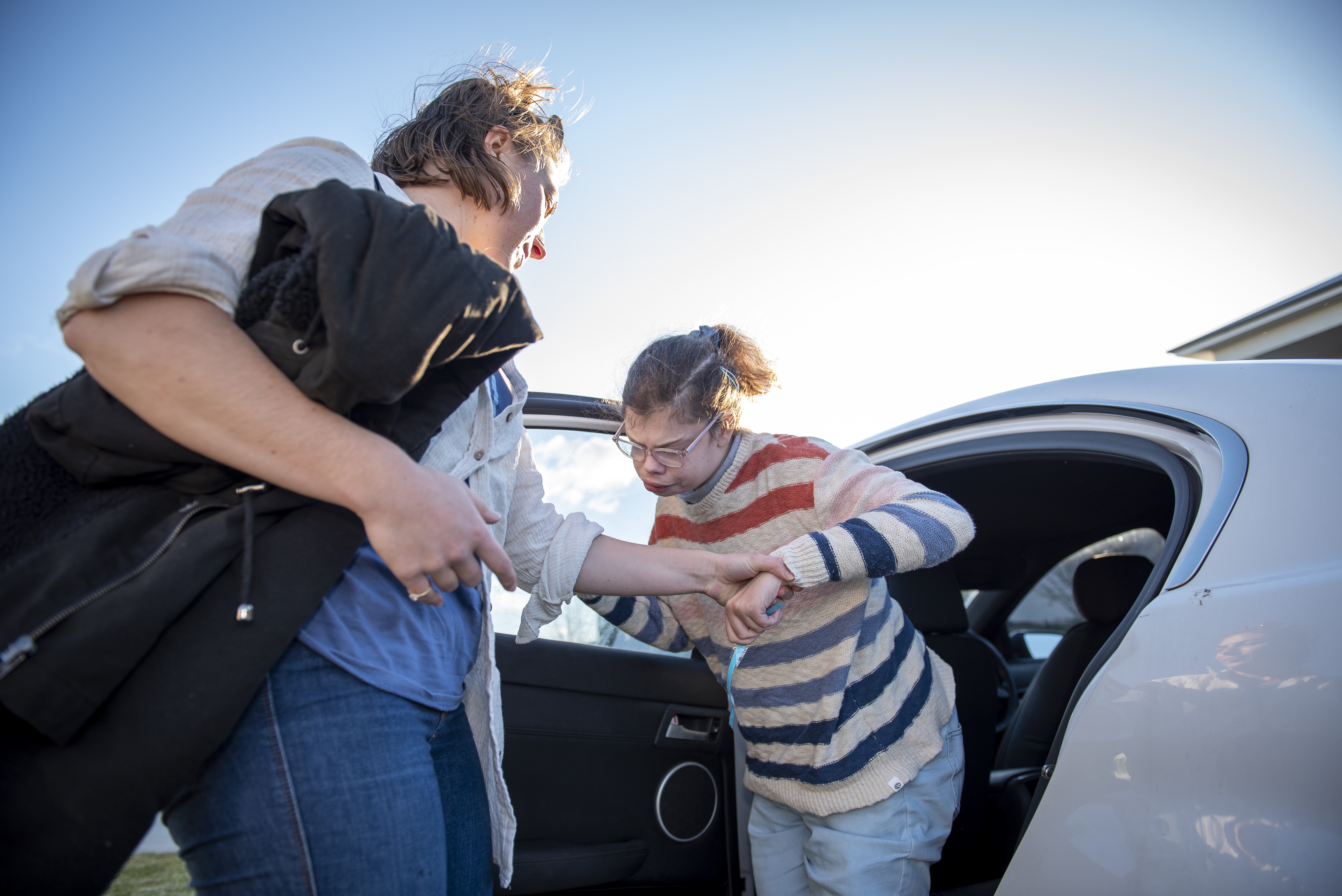 A support worker is assisting a woman getting out of a white car. The Support worker has long brown hair and is wearing a white long sleeved shirt and blue denim jeans. The woman has brown hair tied in a pony-tail and is wearing a white striped jumper and blue jeans.