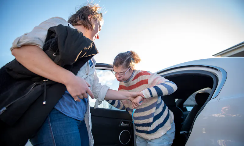 A support worker is assisting a woman getting out of a white car. The Support worker has long brown hair and is wearing a white long sleeved shirt and blue denim jeans. The woman has brown hair tied in a pony-tail and is wearing a white striped jumper and blue jeans.