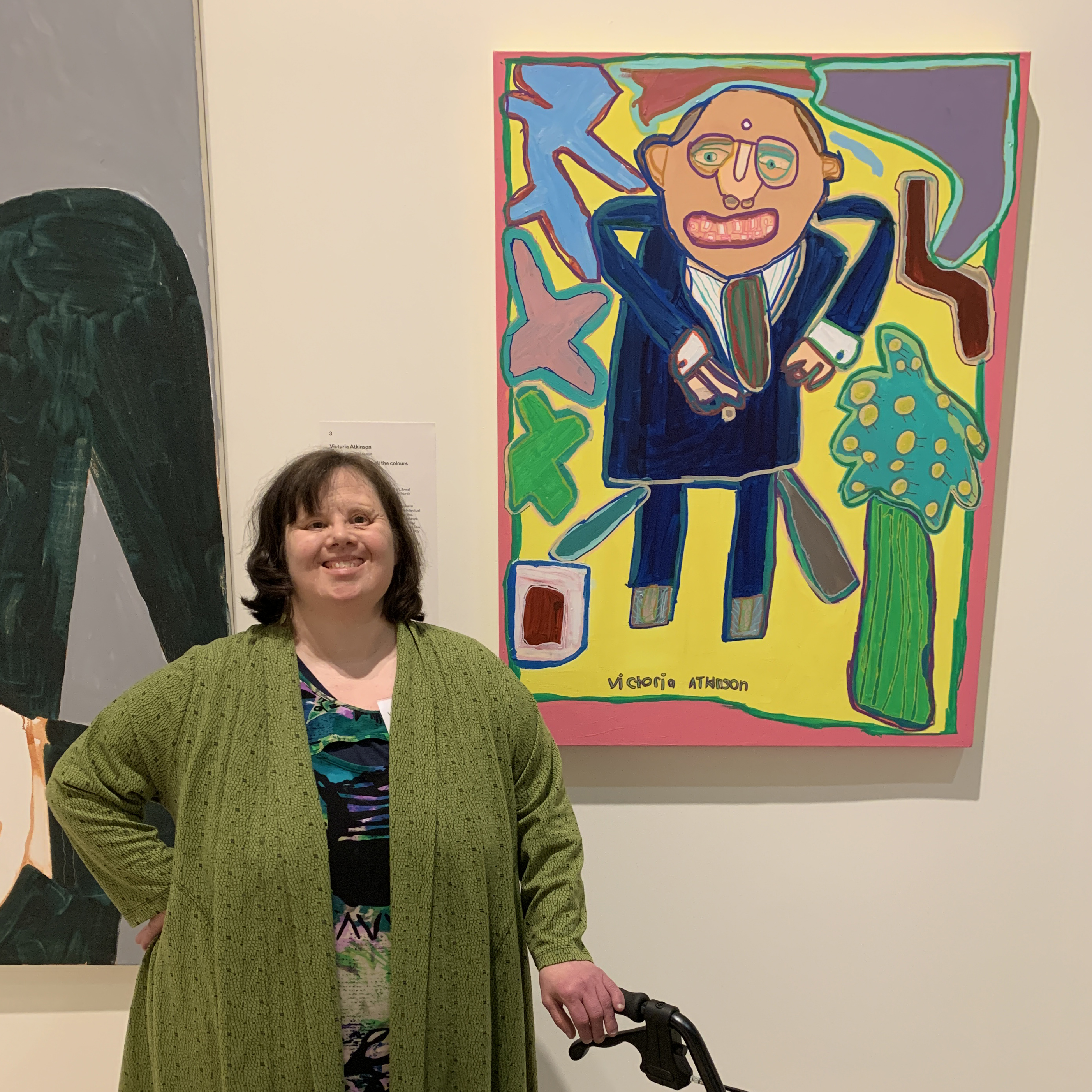 Victoria is wearing a long cardigan and is standing beside her colourful portrait of Trent Zimmerman, which is hanging on a wall. 